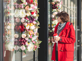 Woman in red duffle coat walks pass florist shop. Woman goes along sunny side of street with cup of coffee to go.