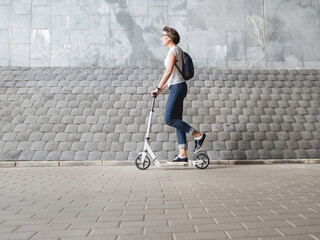 Plakat Woman rides kick scooter. Female in jeans and sneakers moves fast on grey tiled wall background. Healthy lifestyle. Eco-friendly urban transport.