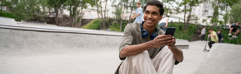 happy african american man with headphones and mobile phone sitting in skate park, banner.