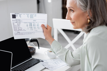 Side view of blurred mature woman holding bills near laptops at home.