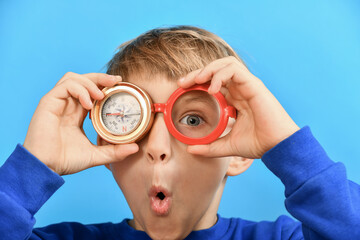 A funny and surprised boy holds a compass in one hand near his eyes and glasses in the other.