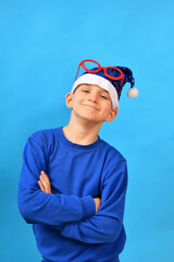 A joyful and smiling boy in a blue Santa Claus hat and red glasses on his head crossed his arms and poses for New Year and Christmas.