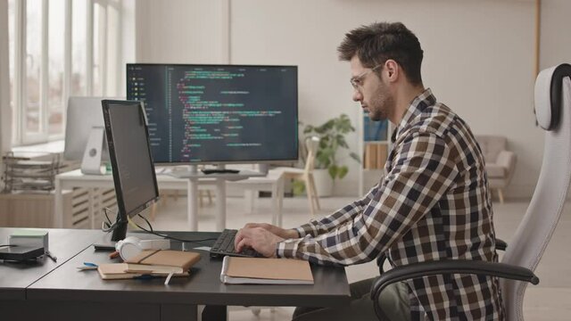 Side-view stab shot of young bearded software engineer or system administrator in casualwear and eyeglasses working with program codes on pc computer in modern office