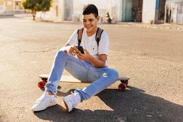 Gordijnen Teenage boy using smartphone sitting on his skateboard in the street © kleberpicui