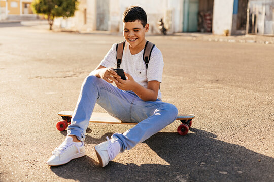 Teenage boy using smartphone sitting on his skateboard in the street