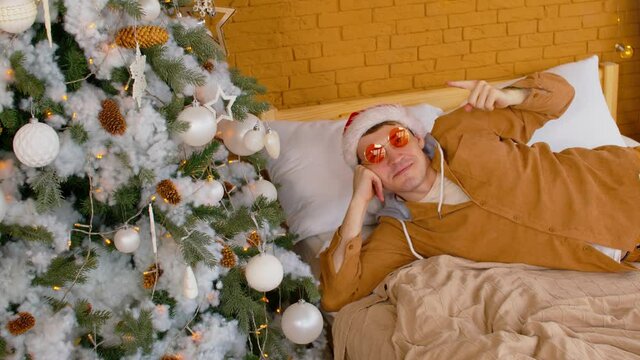Man in santa hat and sunglasses lying on bed near decorated Christmas tree. Stylish male points his finger at coniferous tree with different christmas toys, looking at camera and smiling.