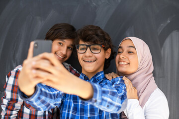 Group of arab teens taking selfie photo on smart phone with black chalkboard in background. Selective focus 