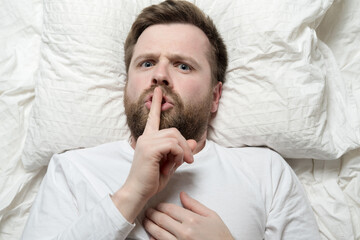Serious man lies in bed and asks to be silent with his finger to his lips. Silence or secret...