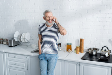 Bearded mature man talking on smartphone near stove in kitchen.