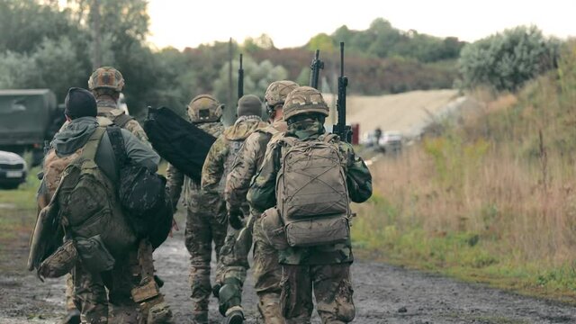 A group of military men returning after completing a task. Soldiers in military uniform with machine guns walk on a stony road with their backs to the cell.