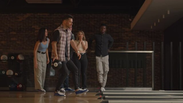 A young Caucasian man makes the final throw with a bowling ball and wins the game with the support and joy of his friends of different nationalities. Rejoice and celebrate the victory