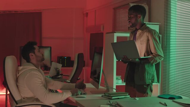 Stab Slowmo Shot Of Two Male Diverse Programmers Having Conversation In Dark Office With Red And Green Lighting