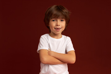 Picture of shy little brunette boy in white t-shirt smiling with embarrassment standing with hands crossed in protection posture. Body language, sign, gestures. Kids children, childhood