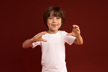 Horizontal picture of adorable male child having cheerful face expression, gesticulating with hands, showing shape of secret subject, playing funny childish game, isolated on red wall