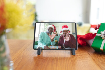 Happy senior african american couple in santa hats waving on tablet video call screen at christmas