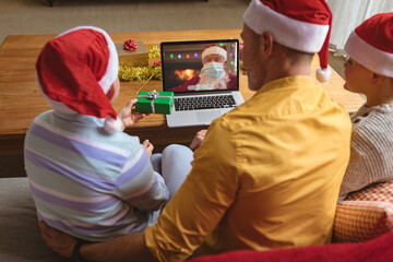 Caucasian father and sons making laptop christmas video call with santa claus in face mask