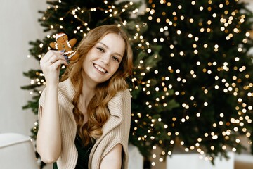 A cheerful beautiful red-haired woman looks into the camera and smiles against the background of a Christmas glowing decorated Christmas tree.