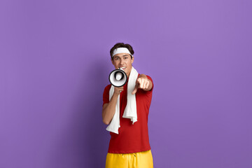 Obraz premium Hey you, start today. Young sportsman is holding loudspeaker and points to camera with finger, wears red t-shirt, yellow shorts, white headband and towel over neck, isolated on violet background.