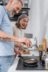 Cheerful woman holding knife near husband cooking pancake in kitchen.
