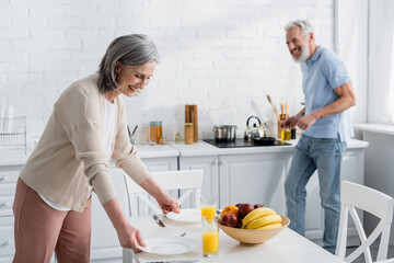 Cheerful mature woman putting plates near orange juice ad blurred husband in kitchen.