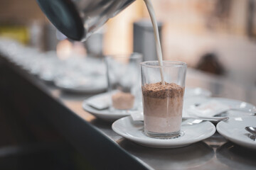
a worker pouring milk into two glasses with chocolate in a cafeteria.