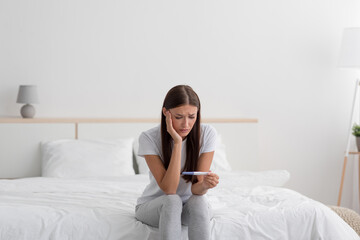 Sad disappointment young european woman hold pregnancy test, sits on bed alone in bedroom