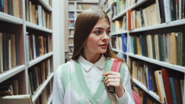 Young woman with long hair wearing white shirt and vest walking among rows of bookcases in library, backpack on her shoulder. Tracking shot of intelligent female looking for literature in labyrinth