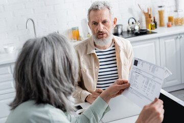 Mature man talking to wife with bills near laptops and smartphone in kitchen.