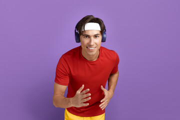 Above shot of running smiling sportsman playing team game, catching ball, listening music, wearing red t-shirt, yellow shorts, blue headphones and white headband, isolated on violet background.