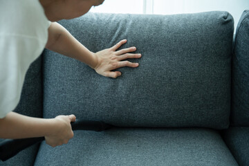 Asian housewife using a wireless vacuum machine to clean a sofa in living room close up. Asian housekeeper vacuuming in living room. Chores and daily housework activities concept.