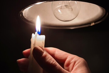 A woman's hand with burning candle in complete darkness looking on a switched off light bulb at...