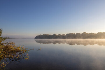 Autumn landscape in the early morning on the river. Misty water surface. Yellow leaves on trees and bushes are illuminated by the rays of the rising sun. Dawn on a cold autumn morning.