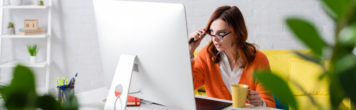 woman with cup of tea touching eyeglasses while working near monitor on blurred foreground, banner