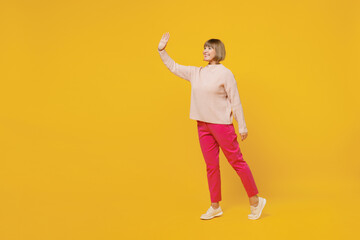 Fototapeta na wymiar Full body side view elderly smiling fun happy caucasian woman 50s with bob haircut wears pink casual knitted sweater walk go waving hand say hello isolated on plain yellow background studio portrait