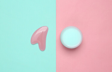 Cream jar and gua sha stone massager on blue pink background. Top view