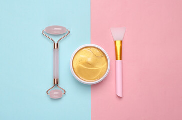 Gold cream jar or patches, facial massage roller on pink blue background. Skin care, lifting face...