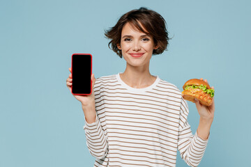 Young smiling happy fun woman 20s in striped shirt hold mobile cell phone with blank screen workspace area burger isolated on plain pastel light blue background studio People lifestyle food concept.