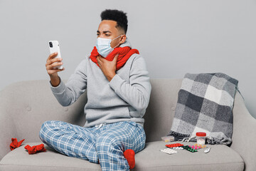 Youngn african man in sweater scarf face mask ppe with sore throat sit on sofa hold use mobile cell phone isolated on plain gray background studio Healthy lifestyle ill sick disease treatment concept