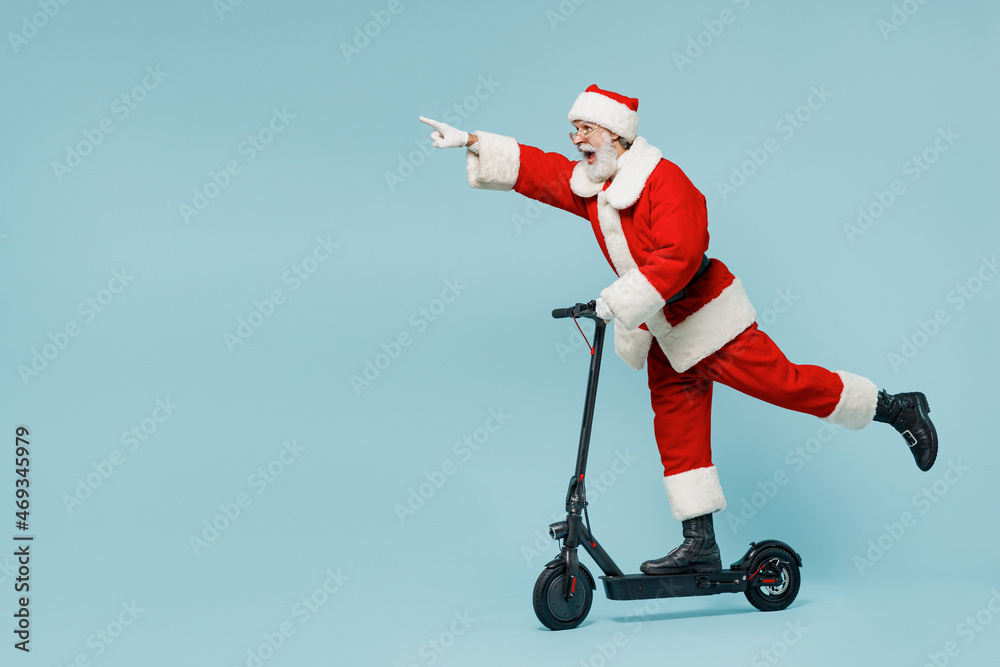 Wall mural Full body fun old Santa Claus man 50s in Christmas hat red suit clothes rides kick scooter point index finger aside isolated on plain blue background studio Happy New Year 2022 merry ho x-mas concept