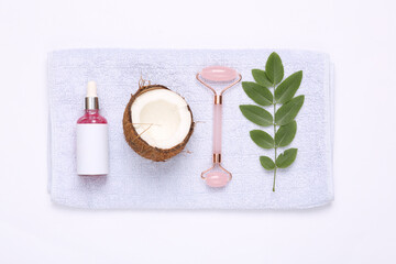 Face roller and serum bottle with green leaves and coconut on towel. Top view. Natural cosmetics