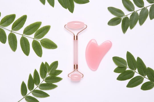 Face roller and gua sha stone massager on white background with green leaves. Top view