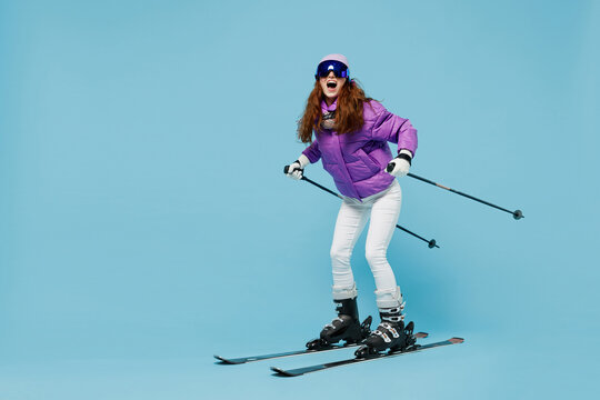 Full body skier shocked happy fun cool woman 20s wearing warm purple padded windbreaker jacket ski goggles mask spend extreme weekend in mountains look camera isolated on plain blue background studio