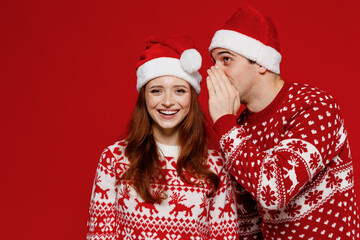 Young happy couple friends two man woman in sweater hat whispering gossip and tell secret behind his hand share news hold face isolated on plain red background Happy New Year 2022 celebration concept.