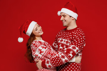 Young side view couple friends two man woman 20s in sweater hat hug cuddle look to each other isolated on plain red background studio Happy New Year 2022 celebration merry ho x-mas holiday concept