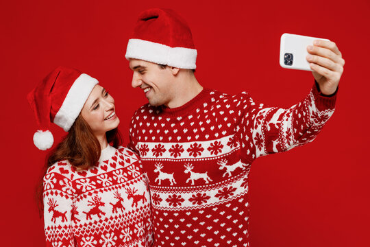 Young couple friends two man woman in sweater hat do selfie shot on mobile cell phone look each other isolated on plain red background. Happy New Year 2022 celebration merry ho x-mas holiday concept