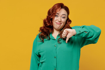 Frowning irritated dissatisfied displeased young ginger chubby overweight woman 20s years old wears green shirt showing thumb down dislike gesture isolated on plain yellow background studio portrait.