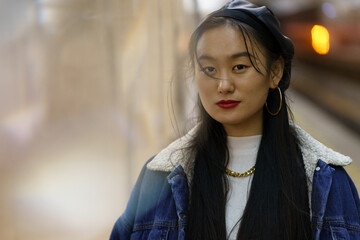 Closeup portrait of millennial asian girl in trendy leather beret and jeans coat wait for train at subway. Young casual chinese female with long black hair. Street style fashion and lifestyle concept