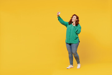 Fototapeta na wymiar Full size body length young ginger chubby overweight woman 20s wear green shirt doing selfie shot on mobile cell phone post photo on social network isolated on plain yellow background studio portrait
