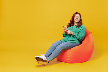 Fototapeta na wymiar Full size body length fun young ginger chubby overweight woman 20s wear green shirt sit in bag chair hold in hand use mobile cell phone look camera isolated on plain yellow background studio portrait
