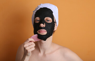 Beautiful woman with purifying black mask on her face and towel on head. Girl massages face gua sha stone. Orange background. Facial skin care